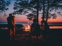 Group of young people near camp fire telling stories near the fire with wood, flames in the nature at night near lake.