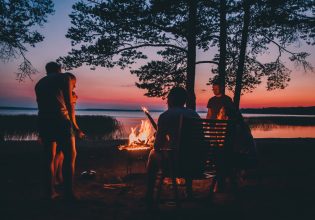Group of young people near camp fire telling stories near the fire with wood, flames in the nature at night near lake.